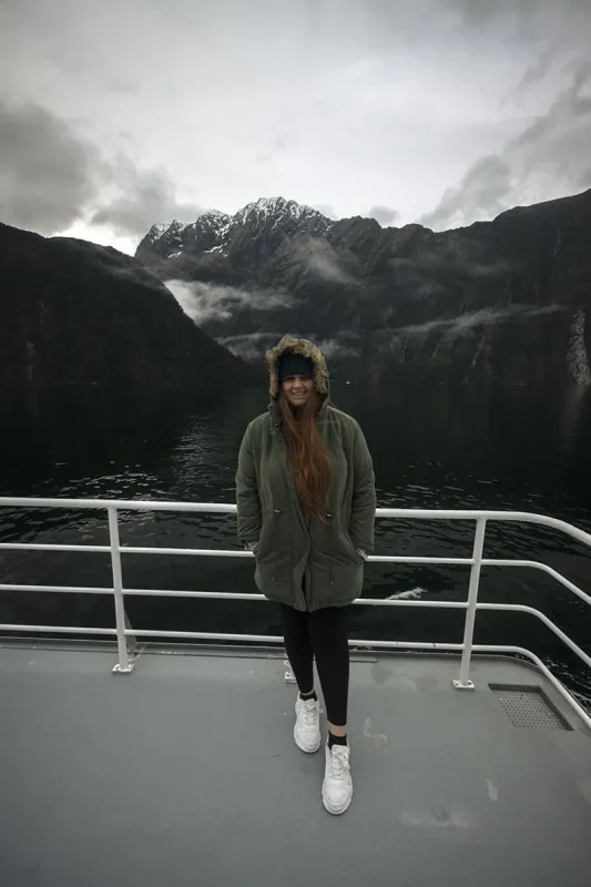 Milford Sound In Winter: Is It Still Worth The Hype? 5 tasha amy stands on the deck of a boat, smiling at the camera, wrapped in a green parka with a fur-lined hood. The dark, tranquil waters of Milford Sound surround the boat, and towering, snow-capped mountains rise majestically in the background. The overcast sky and mist add to the serene and cool ambiance of winter in Milford Sound.