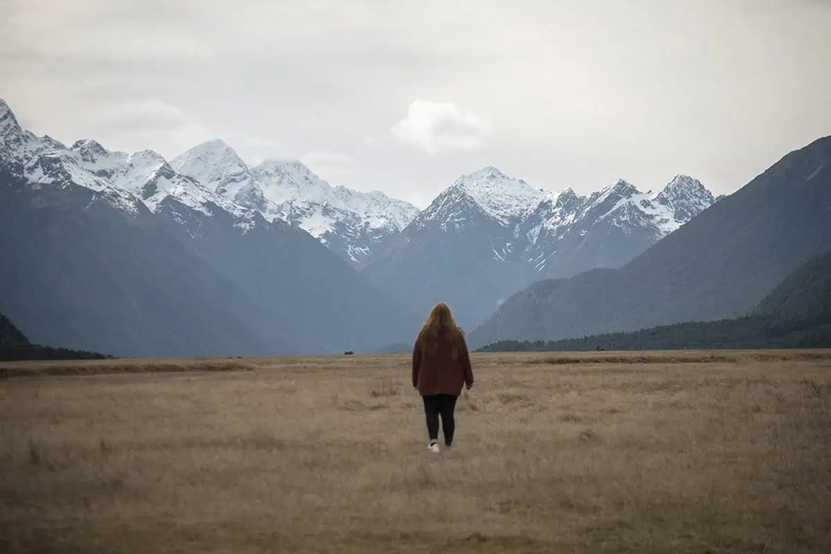 Milford Sound In Winter: Is It Still Worth The Hype? 7 tasha amy walks alone across a vast, grassy field towards snow-capped mountains in the distance. The overcast sky and the expansive, open landscape create a sense of solitude and reflection, typical of winter in Milford Sound.
