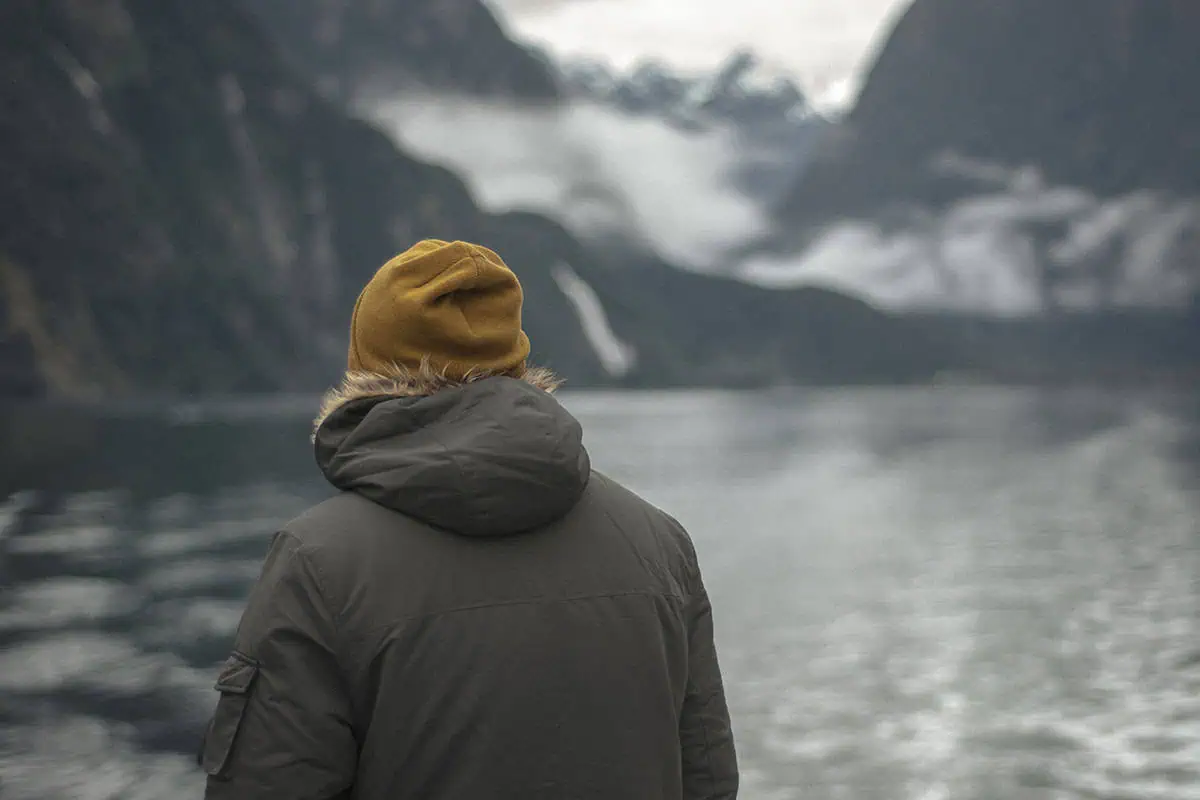 Milford Sound In Winter: Is It Still Worth The Hype? 12 A person with their back to the camera stands on the edge of a body of water, wearing a mustard-yellow beanie and a dark grey parka with a fur-lined hood. The overcast sky and mist shroud the towering mountains in the background, creating a serene and contemplative atmosphere typical of winter in Milford Sound.
