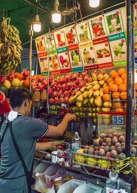 120+ Would You Rather Travel Questions For Ultimate Wanderers 14 The stall features stacks of mangoes, oranges, dragon fruit, and bananas with a large drink menu hanging overhead and blenders on the counter in bangkok
