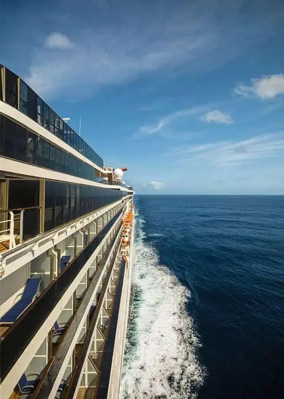 120+ Would You Rather Travel Questions For Ultimate Wanderers 5 The ship leaves a white trail of wake behind it under a clear sky, with multiple decks and balconies visible along its length.