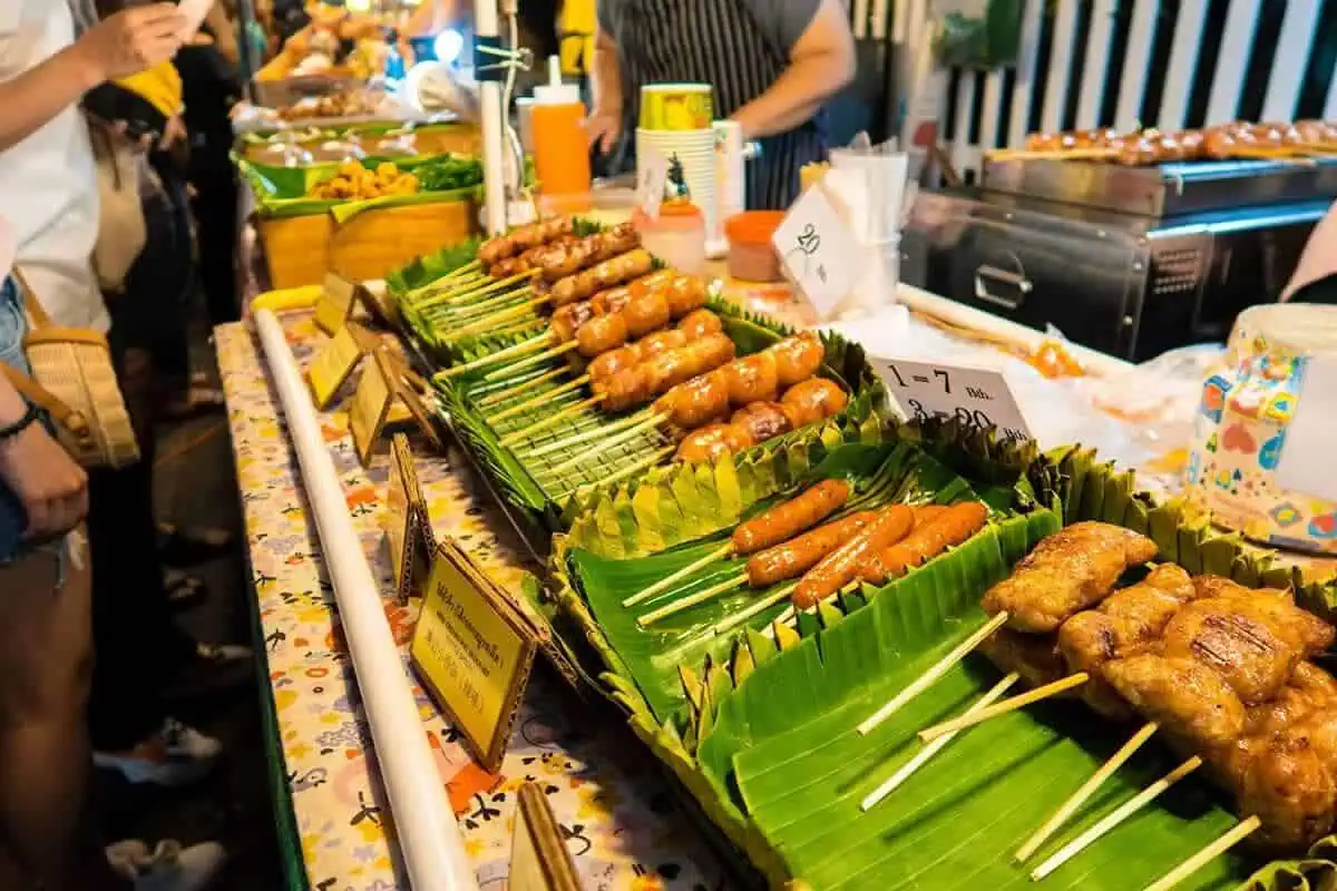 120+ Would You Rather Travel Questions For Ultimate Wanderers 2 Vendors serve various skewered sausages and meats to customers at a vibrant market stall, with handwritten signs showing prices in thailand