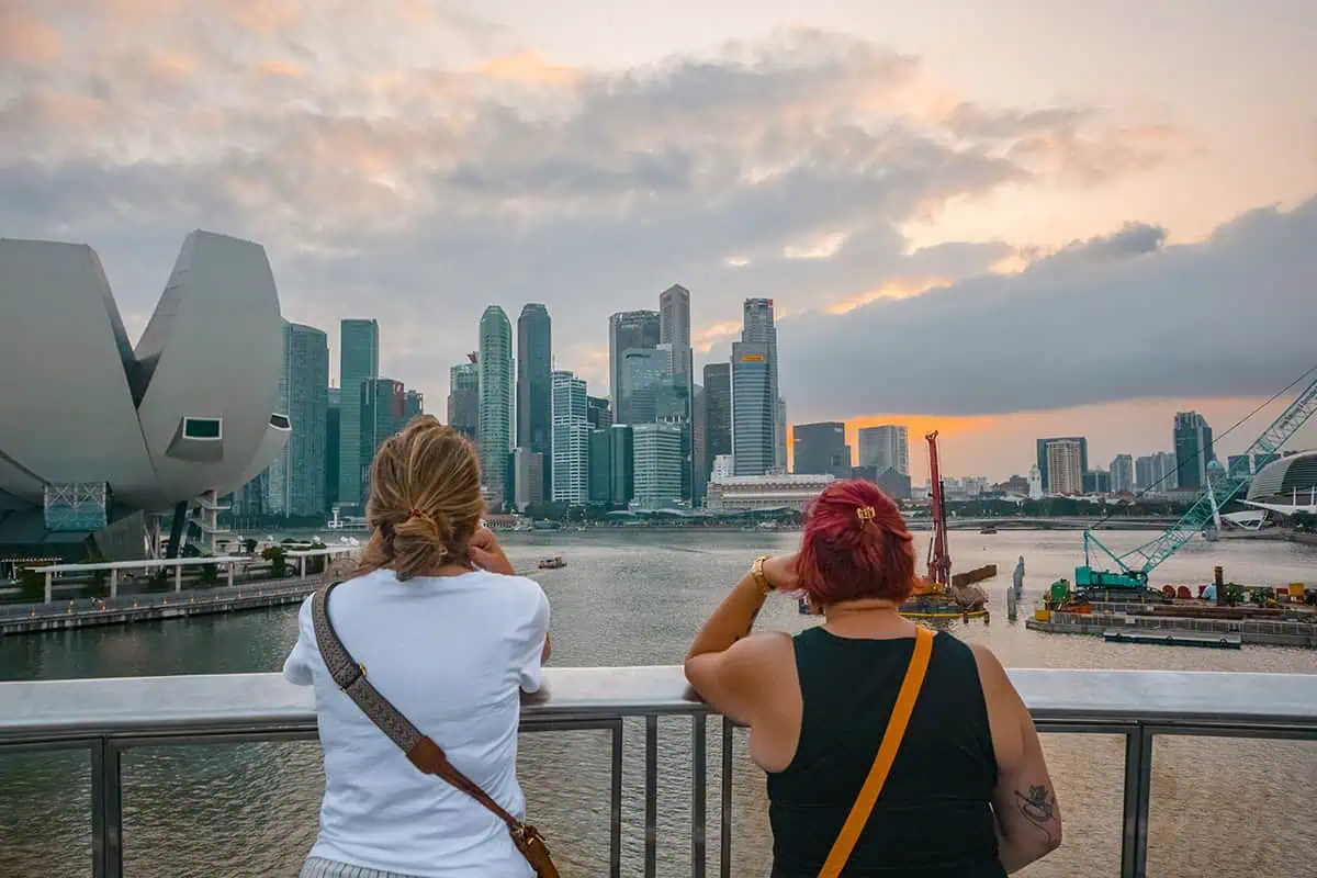120+ Would You Rather Travel Questions For Ultimate Wanderers 3 Looking across Marina Bay in Singapore, two ladies look at the modern skyscrapers with sunset overhead and boats below.