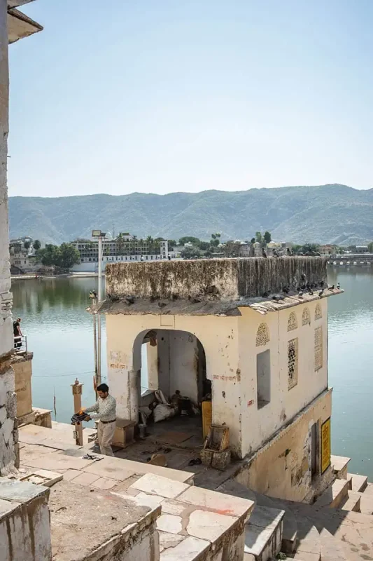 120+ Would You Rather Travel Questions For Ultimate Wanderers 12 A local man tends to tasks beside a lakeside temple in Pushkar, India, with still waters reflecting surrounding buildings and mountains.